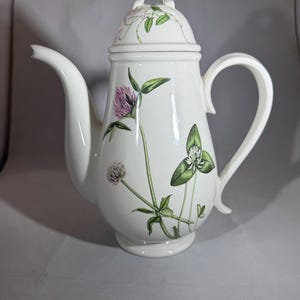 May include: White ceramic teapot with a lid, decorated with a floral pattern of pink and green clovers. The teapot has a curved spout and a large handle. The lid has a small loop handle. The design is a botanical illustration.
