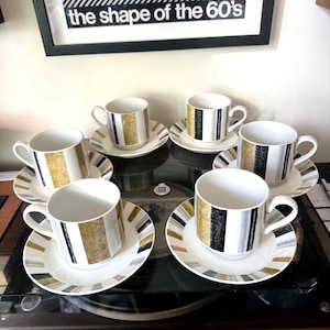 Op de afbeelding: Een set van vier witte keramische theekopjes met bijpassende schoteltjes, voorzien van verticale strepen in metallic goud, zilver en zwart. Elk kopje heeft een gebogen oortje en de schoteltjes tonen een stralend patroon van afwisselende gekleurde strepen.