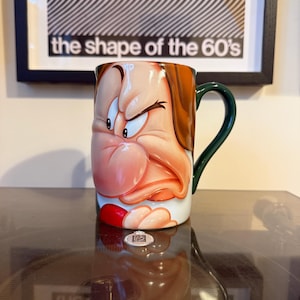 Può includere: Una tazza in ceramica con il volto di un personaggio dei cartoni animati con un'espressione arrabbiata. La tazza ha un manico verde e un interno bianco. Il personaggio ha i capelli castani, un colletto rosso e un naso rosa. La tazza è su una superficie riflettente.