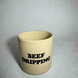 May include: A cream-coloured ceramic container with the words "BEEF DRIPPING" in black lettering. The cylindrical container has a smooth finish and a classic design, suitable for kitchen use.
