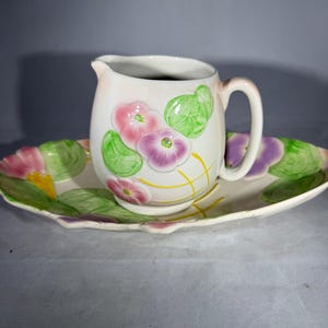 May include: A cream-colored ceramic creamer and serving platter set. The creamer and platter are decorated with pink, purple, and green floral designs. The platter has a scalloped edge.