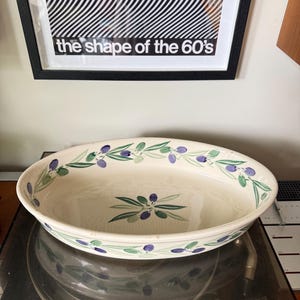 May include: An oval ceramic serving bowl with a cream-colored base, featuring a hand-painted pattern of green leaves and purple olives around the inner rim and a central cluster of olives and leaves at the bottom of the interior basin.