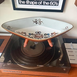 May include: A diamond-shaped ceramic bowl with three small textured feet. The interior features a white base with brown, blue, and black scrollwork patterns. The exterior rim is a light teal color with brown accents, and the outer base is brown.