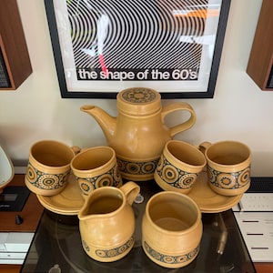 May include: A tan stoneware tea set featuring a teapot with a lid, four mugs with matching saucers, a creamer, and a sugar bowl. Each piece is decorated with a dark green and black circular geometric pattern around the center.