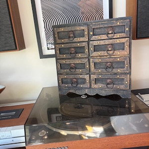 May include: A small wooden chest with eight drawers, each featuring a dark finish, gold-toned metal studs, and a circular metal pull handle. The chest is positioned on top of a Beogram 1000 record player with a wooden base and a dark plastic cover.