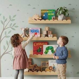 May include: Four light-colored wooden wall shelves with scalloped edges, holding various children's books including Goodnight Moon, Mama, Corduroy, and The Very Hungry Caterpillar, along with wooden blocks, a toy truck, and a small potted plant.