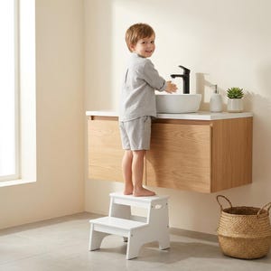 May include: A white two-step wooden stool with side handles, positioned in front of a wall-mounted light wood vanity cabinet featuring a white countertop, a white vessel sink, a black faucet, and a white soap dispenser.