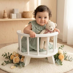 May include: A baby with dark curly hair wears a sage green sleeveless dress with a ruffled collar and small floral embroidery. The baby holds a cream-colored knitted teddy bear with a dusty pink ribbon around its neck while sitting in a white chair.