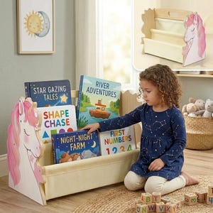 May include: A wooden children's bookshelf featuring a pink unicorn design on the side panels and fabric slings for book storage. The shelf holds books titled Star Gazing Tales, Shape Chase, River Adventures, Night-Night Farm, and First Numbers.