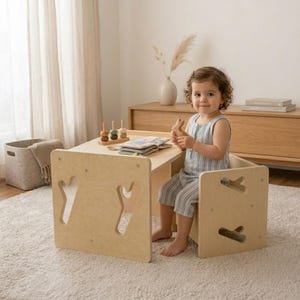 May include: A light-colored wooden table and chair set with Y-shaped cutouts on the sides. The table holds a small wooden toy and an open book. The furniture features a smooth, natural finish and a modular design suitable for a child's play area.