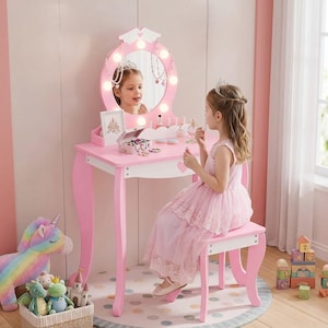 May include: A pink and white wooden vanity table with a round mirror featuring integrated lights. The set includes a matching stool, a small storage box, and a frame. The vanity has curved legs and a scalloped edge design on the tabletop surface.