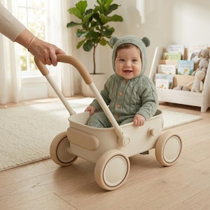 May include: A baby sits inside a cream-colored toy wagon with four wheels and a handle. The baby wears a sage green cable-knit onesie with a matching bear-ear hood. The wagon features a smooth, rounded design with tan accents on the wheels and handle.