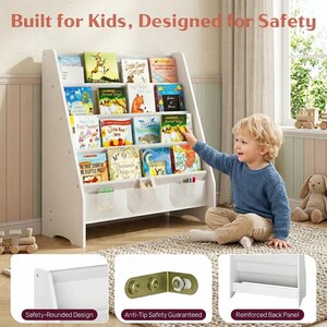 May include: White wooden bookshelf filled with children's books. The bookshelf has a slanted design and includes fabric storage pockets. The text "Built for Kids, Designed for Safety" is at the top. A young child is reaching for a book.