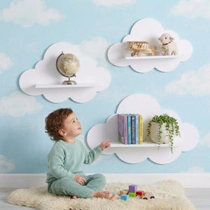 May include: Three white cloud-shaped wall shelves mounted on a blue sky wallpaper. The top left shelf holds a globe, the top right shelf holds a wooden toy airplane and a plush lamb, and the bottom shelf holds a row of books and a potted plant.