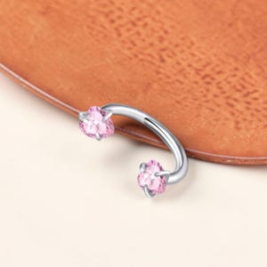 Puede incluir: Una joya de piercing curvada, de estilo barra, de color plateado, con gemas rosas en forma de corazón en cada extremo. La joya está colocada sobre una superficie marrón.
