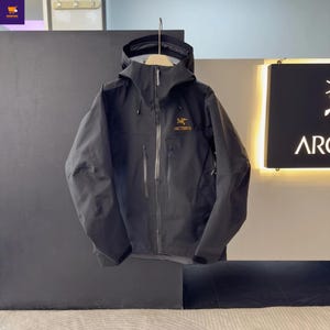 May include: A black hooded jacket with the brand name "Arc'teryx" in gold lettering. The jacket has a zipper closure, multiple pockets, and a drawstring hood. The jacket is hanging on a wooden hanger.