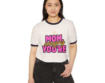 May include: A white ringer t-shirt with dark blue trim on the collar and sleeves. The front features a gold crown icon above the text "MOM. amazing YOU'RE" printed in bold pink and yellow lettering. The shirt is paired with dark grey denim jeans.