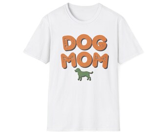 May include: A white short-sleeved t-shirt featuring the text DOG MOM in large, orange, distressed-style block letters. Below the text is a small, solid green silhouette of a dog standing in profile, centred on the chest of the garment.