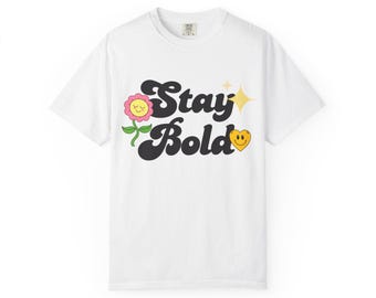 May include: A white short-sleeved t-shirt featuring the black text Stay Bold in a wavy font. The design includes a pink flower with a smiling face, a yellow four-pointed star, and a yellow heart with a smiling face on the right side of the text.