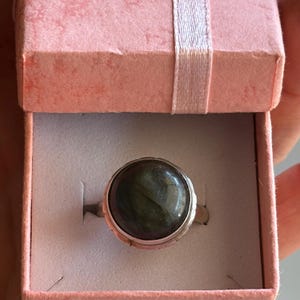 May include: A silver ring with a large, round, dark green gemstone, presented in a pink gift box with a white ribbon. The ring's setting has a decorative edge.