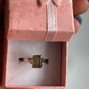 May include: A silver ring with a large, emerald-cut, olive-green gemstone is presented in a pink gift box. The ring features detailed band work. The box is open, showing the ring against a white interior.
