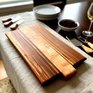 Peut inclure: Planche à découper en bois avec une poignée, présentant un mélange de veines de bois claires et foncées. La planche est rectangulaire avec une poignée arrondie. Couteaux à fromage, assiettes et un bol de baies sont à proximité.