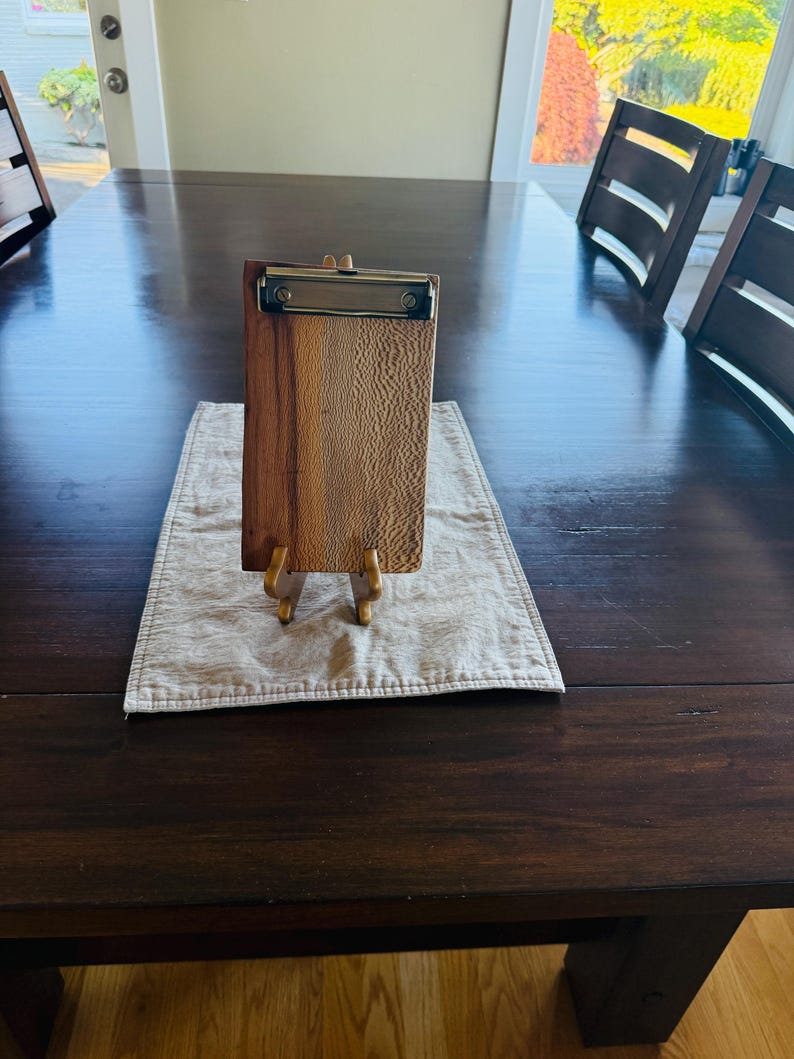 May include: A rectangular wooden clipboard featuring a distinct two-tone grain pattern with a smooth, light-colored textured section on the right and a darker, solid wood section on the left, topped with a metallic silver-toned spring clip.