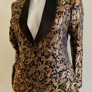 Puede incluir: Una chaqueta tipo blazer con base negra y un patrón de cachemira en tonos oro y cobre metalizados. La prenda incluye un cuello chal negro liso, mangas largas, un cierre frontal de un solo botón y un bolsillo ribeteado en el lado izquierdo.