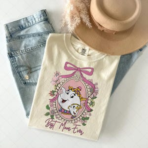 May include: Cream-colored t-shirt featuring a cartoon illustration of Mrs. Potts and Chip from Beauty and the Beast, framed in pink with a pink bow and the text "Best Mom Ever." The shirt is paired with light-wash jeans and a tan hat.