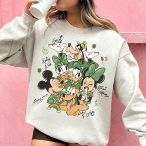 Puede incluir: Una sudadera gris claro con un gráfico colorido de personajes de Disney, incluyendo Mickey Mouse, Minnie Mouse, Donald Duck, Daisy Duck, Goofy y Pluto. El diseño incluye tréboles y los nombres de los personajes.