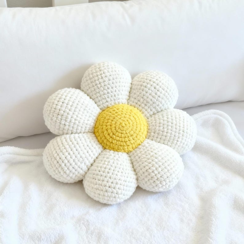May include: A daisy-shaped pillow with white petals and a yellow center. The pillow is made of a textured, crocheted material and rests on a white blanket. The design is a cheerful, decorative accent for a bed or sofa.