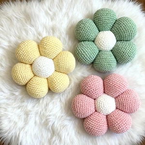May include: Three handmade crochet flower pillows in pastel colors: yellow, green, and pink. Each flower has a white center and is set against a fluffy white background. These decorative pillows add a touch of whimsy to any room.