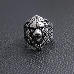 May include: A silver-colored ring featuring a detailed lion's head design. The ring has a textured surface and is set against a dark background. The lion's mane and facial features are intricately carved.