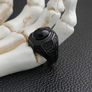 May include: A black ring with a large, oval, black stone set in a decorative, geometric design. The ring is displayed on a white, skeletal hand against a dark, textured background.