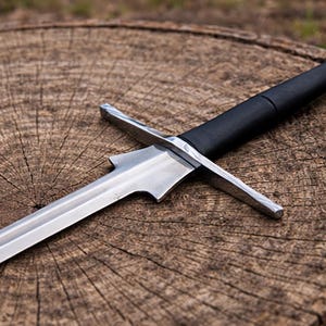 Può includere: Una spada medievale con una lama e una guardia incrociata in acciaio lucido. L'impugnatura è avvolta in materiale nero e il pomo è argentato. La spada poggia su una superficie di legno circolare e usurata.
