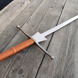 Puede incluir: Una espada larga y plateada con un mango envuelto en cuero marrón y una guarda cruzada. La hoja es recta y se estrecha hasta un punto. La espada descansa sobre una superficie de madera envejecida.