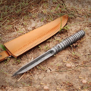 May include: A Damascus steel knife with a patterned blade and a textured, dark gray handle. The knife rests beside a tan leather sheath with decorative stitching. The knife is designed for outdoor use.