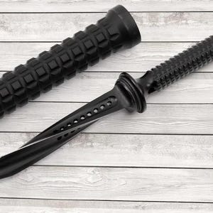 Pode incluir: Uma adaga preta com uma pega texturizada e uma lâmina afiada e curva. A pega tem um padrão de diamante e a lâmina apresenta uma série de orifícios. A adaga está ao lado de um objeto cilíndrico preto com um padrão texturizado semelhante.