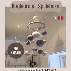 Puede incluir: Móvil colgante con esferas de ganchillo en varios tamaños y colores neutros. El texto "Kugleuro m. Spilleboks" está en la parte superior, con "PDF Pattern" y "Pattern available in US/DE/DA". La bandera danesa es visible.