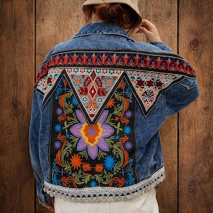 Może przedstawiać: Kurtka dżinsowa z kolorowymi haftowanymi wzorami kwiatowymi i geometrycznymi na plecach. Haft zawiera odcienie czerwieni, pomarańczy, błękitu i fioletu. Kurtka ma białe koronkowe wykończenie wzdłuż dołu.