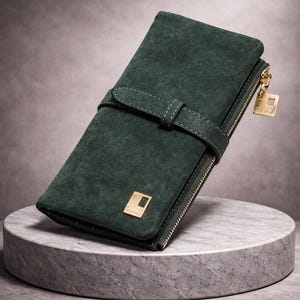 Puede incluir: Una cartera de ante verde oscuro con cremallera y cierre de hebilla dorados. La cartera tiene forma rectangular con una correa y un pequeño emblema cuadrado dorado. El tirador de la cremallera tiene un dije cuadrado dorado.