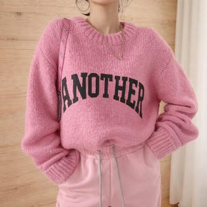 May include: A pink knitted jumper with the word "ANOTHER" in black block letters across the front. The jumper has a crew neck and ribbed cuffs. The model is wearing pink tracksuit bottoms.