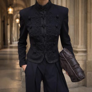 Peut inclure: Veste noire de style militaire avec des fermetures à boucle décoratives et un col montant. La veste est associée à un pantalon sombre et à une pochette en cuir marron. L'ensemble est sophistiqué, adapté aux occasions formelles.