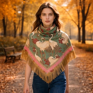 May include: A green scarf with a crane and floral pattern, pink border, and tan fringe. The scarf is worn around the neck and shoulders. The background is a park with trees and fall foliage.