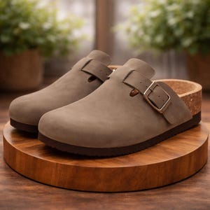 Può includere: Un paio di zoccoli color talpa con chiusura a fibbia. Gli zoccoli hanno una suola in sughero e una suola in gomma marrone scuro. Le scarpe sono esposte su un supporto di legno a forma di cuore.