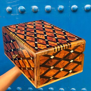 May include: A rectangular wooden box featuring a complex diamond-patterned inlay of light and dark wood grains, accented with thin black strips and small metallic rectangular details. The box includes a small gold-colored metal flower-shaped knob.