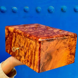 Peut inclure: Une boîte rectangulaire en bois avec une finition polie très brillante, présentant un motif de grain de bois ronceux tourbillonnant dans des tons ambrés et brun foncé. Une petite clé en métal doré est insérée dans la serrure frontale.