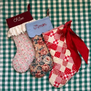 Op de afbeelding: Vier kerstsokken met namen. Eén is bordeauxrood met de naam "Chloe" in wit geborduurd. Een andere is lichtblauw met "Timon" in rood geborduurd. De andere twee hebben bloemen- en patchworkpatronen.