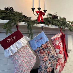May include: Three quilted Christmas stockings hanging in a row. The first is maroon with white lace trim and the name Chloe. The second is light blue with a scalloped edge and the name Timon. The third is a red and white patchwork pattern.
