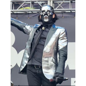 May include: A performer in a silver metallic blazer, black shirt, and black pants, wearing a skull mask. The blazer has a shiny, reflective surface. The performer is holding a microphone.